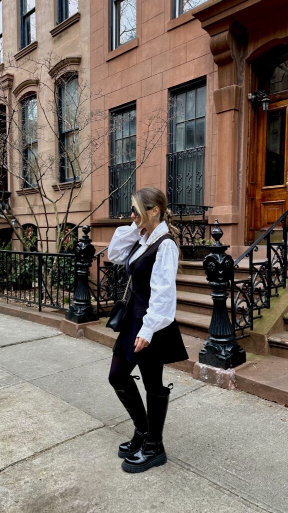 White shirt, long black vest, black tights and chunky black knee-high boots on a city street